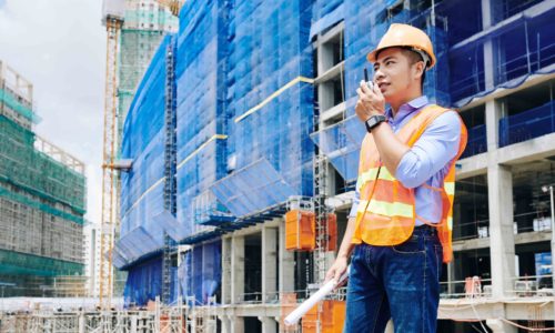 Asian contractor in bright orange vest using walkie-talkie for communication with coworkers at construction site